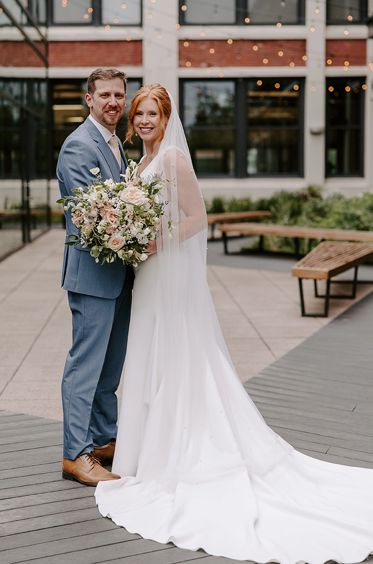 Groom embraces bride in a sleeveless crepe wedding dress with a plunging neck and veil, holding a bouquet in a courtyard.