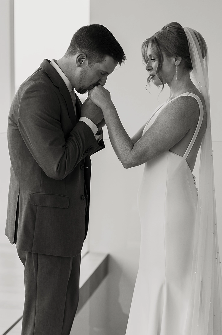 Groom kisses bride's hand, wearing a crepe fitted gown with princess seams and a veil, shown in black and white