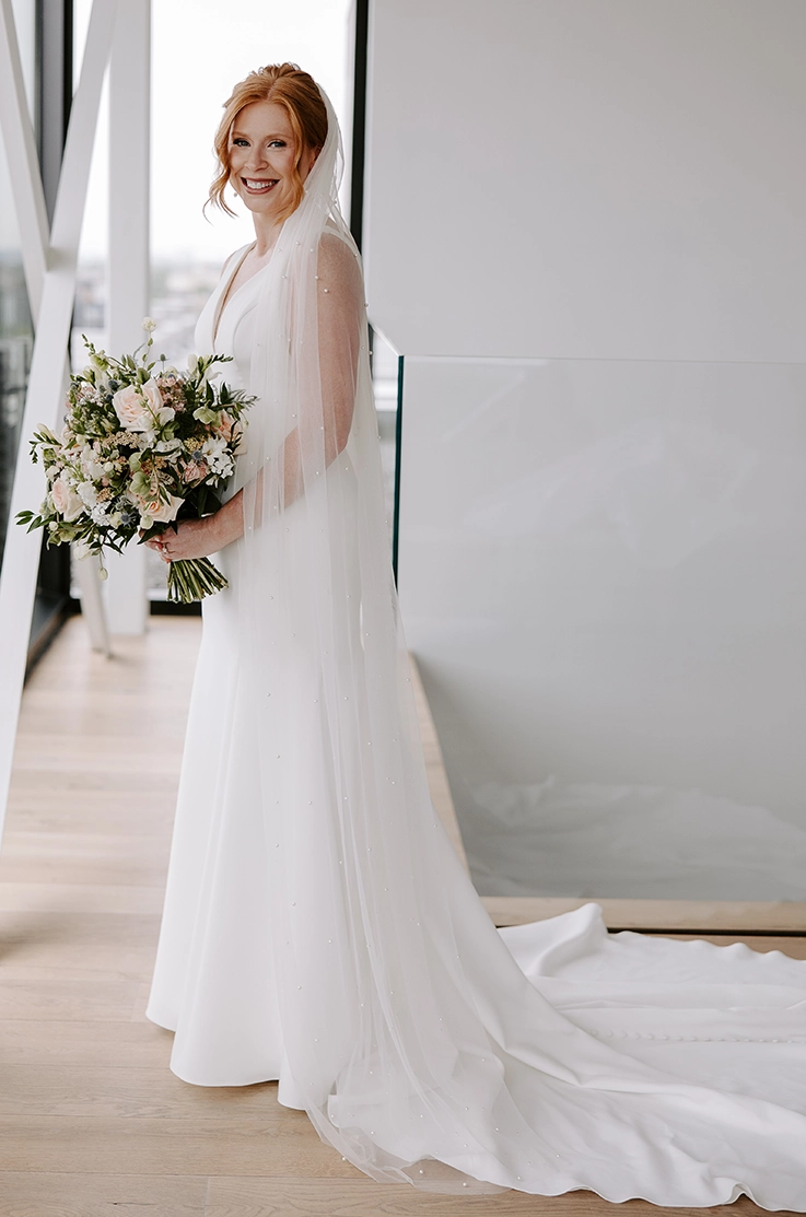 Smiling bride poses in a V-neck crepe gown with princess seams and a long veil, holding a bouquet indoors.