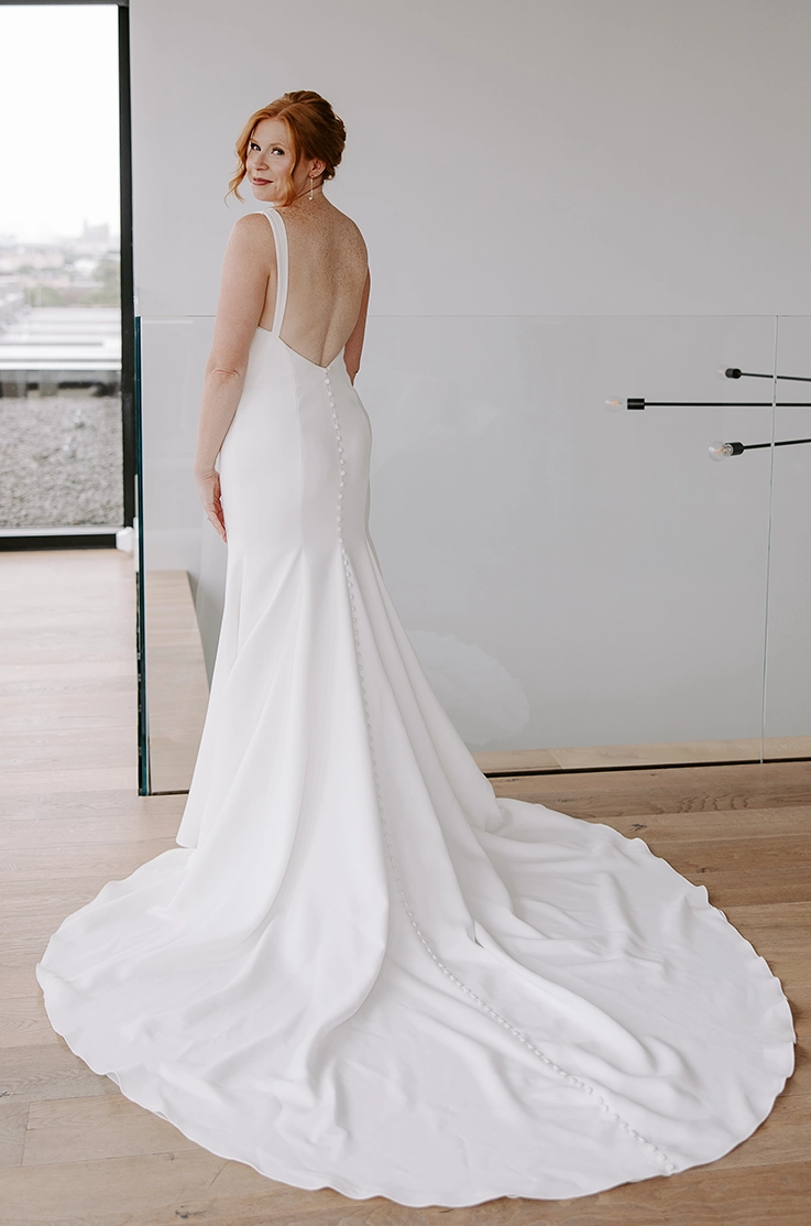 Back of bride in a fit and flare crepe gown with a low buttoned back by a large window and railing. 