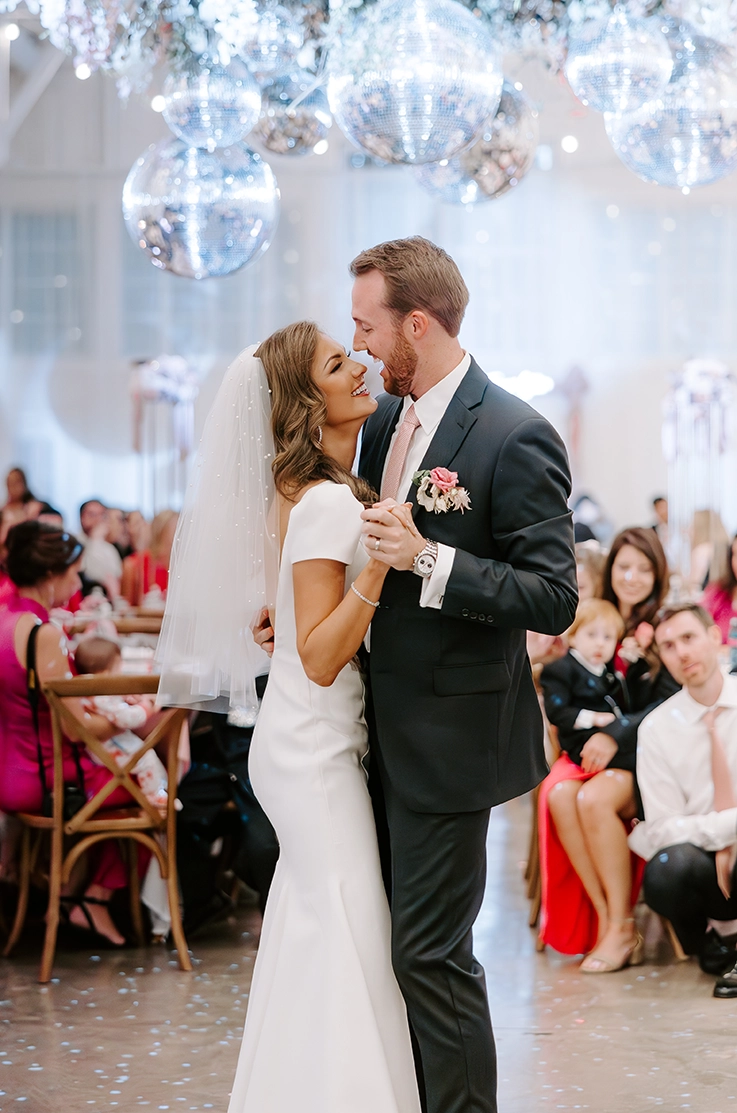Groom dances with his bride under disco balls, in a crepe princess seamed gown with a low back, cap sleeves and a veil. 
