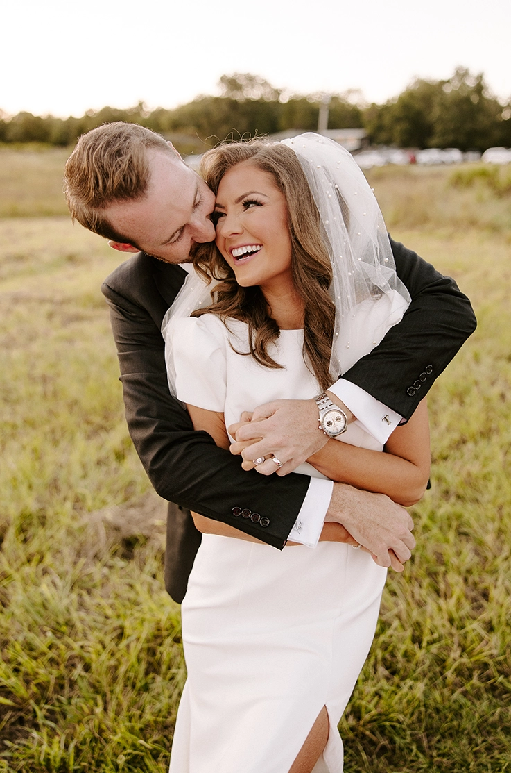 Groom embraces his bride in a crepe bateau neck wedding dress with a skirt slit and pearl veil, in an open meadow. 