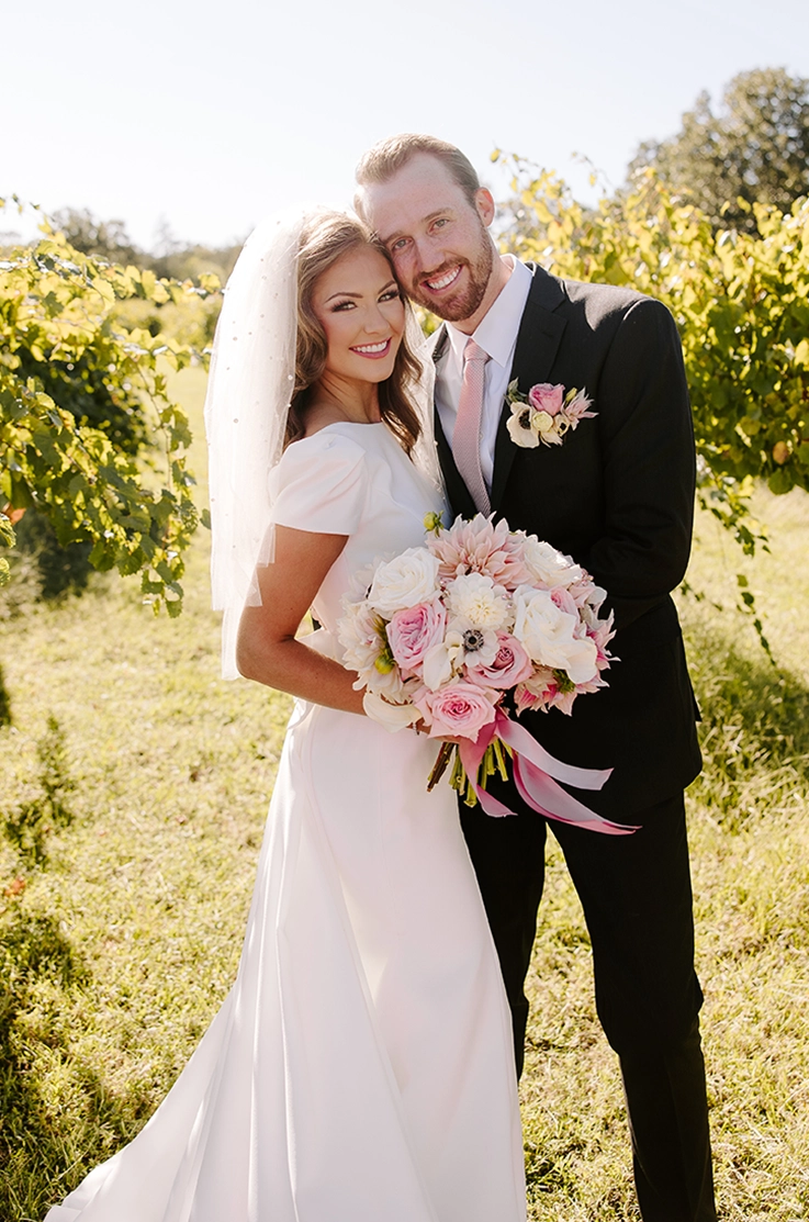 Bride in a crepe fit and flare gown with cap sleeves and a pearl-edge veil, holding a bouquet in a vineyard next to groom.