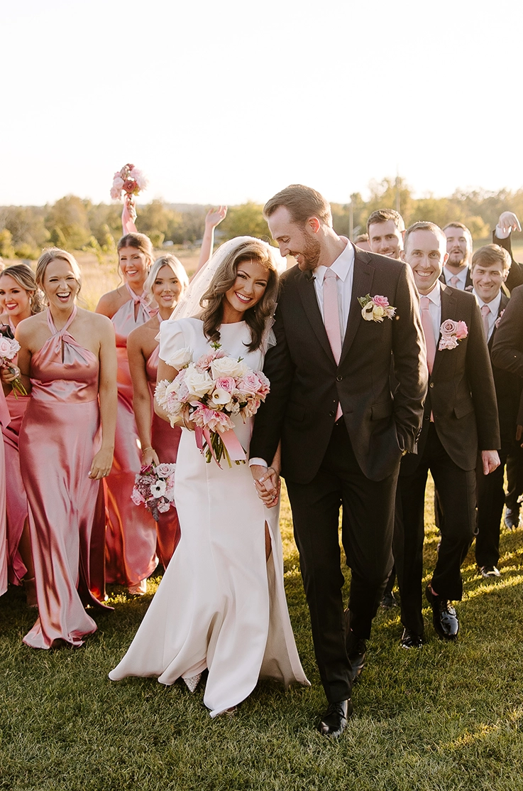 Groom looks at bride in a crepe cap sleeve gown with a skirt slit and veil, holding a blush bouquet in front of bridal party.