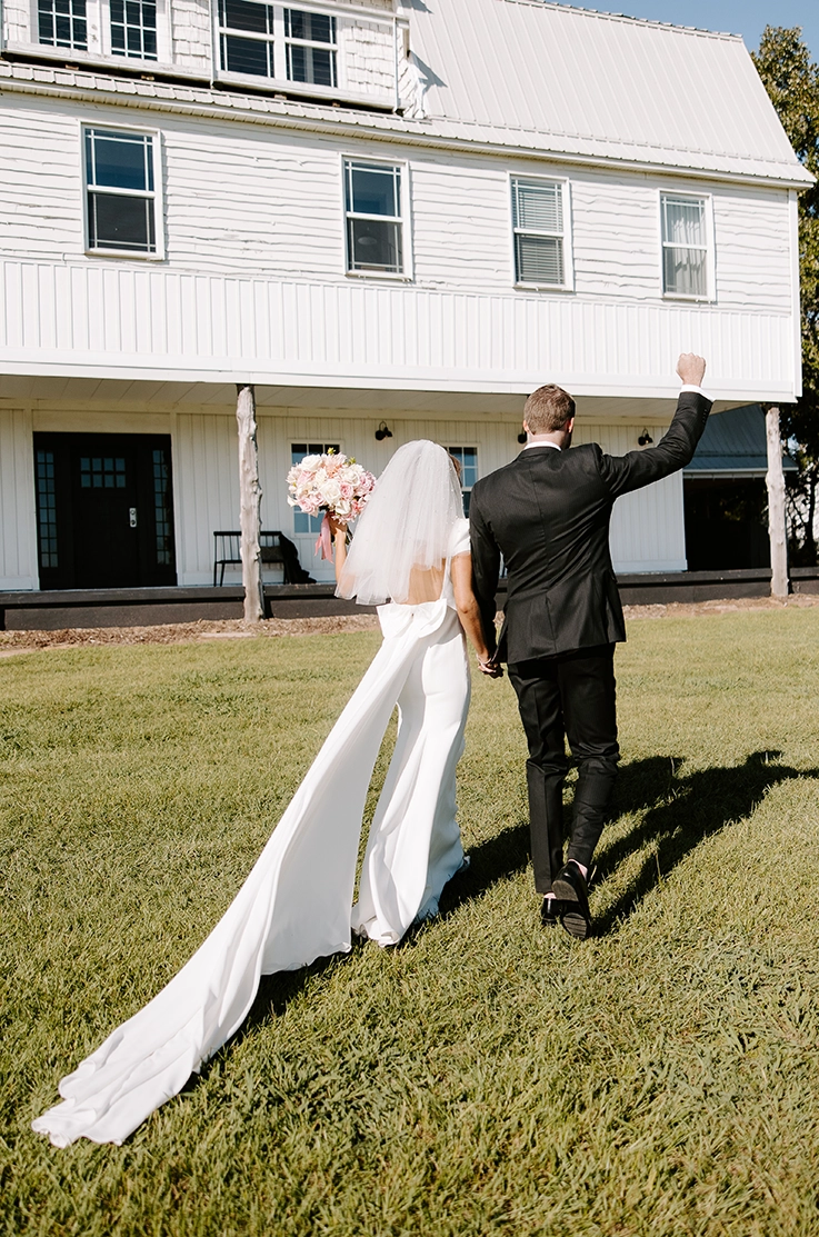  Groom walks alongside bride in a low back fitted gown with a bow, veil and flowing train, holding a bouquet outside the venue. 
