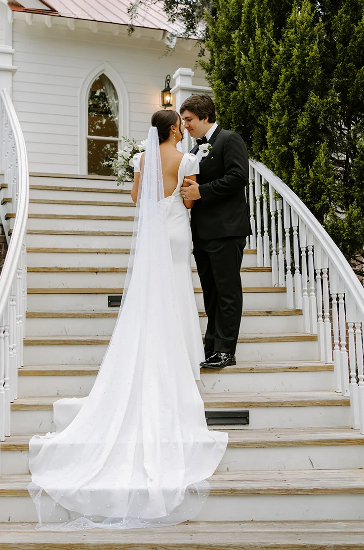 Groom poses with bride in a crepe fit and flare wedding dress with a low back and a veil, standing on stairs outdoors.