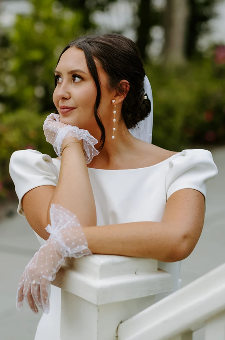 Close-up of bride in a bateau neck crepe gown with gathered cap sleeves, gloves and a veil, leaning on a railing.