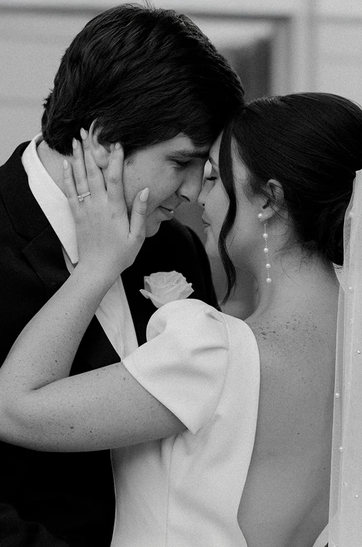 Close-up of groom facing bride in a crepe cap sleeve gown with a low open back and pearl veil, in black and white.