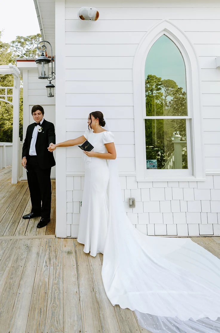 Bride in a bateau neck crepe gown with fit and flare skirt and a pearl-edge veil, posing with groom outside a white chapel.