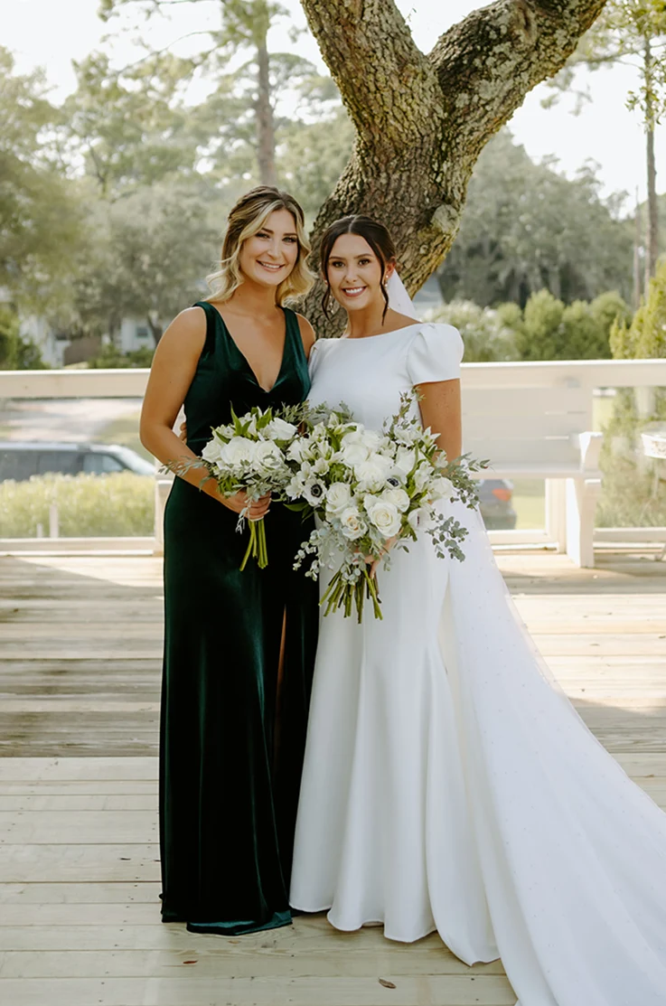 Bride in a puff sleeve crepe gown with a bateau neck and a veil, holds a bouquet with a bridesmaid under a tree in the sun. 