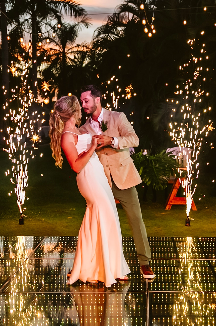 Groom dances with his bride in a crepe fit and flare wedding dress, in front of sparklers and palm trees at sunset.