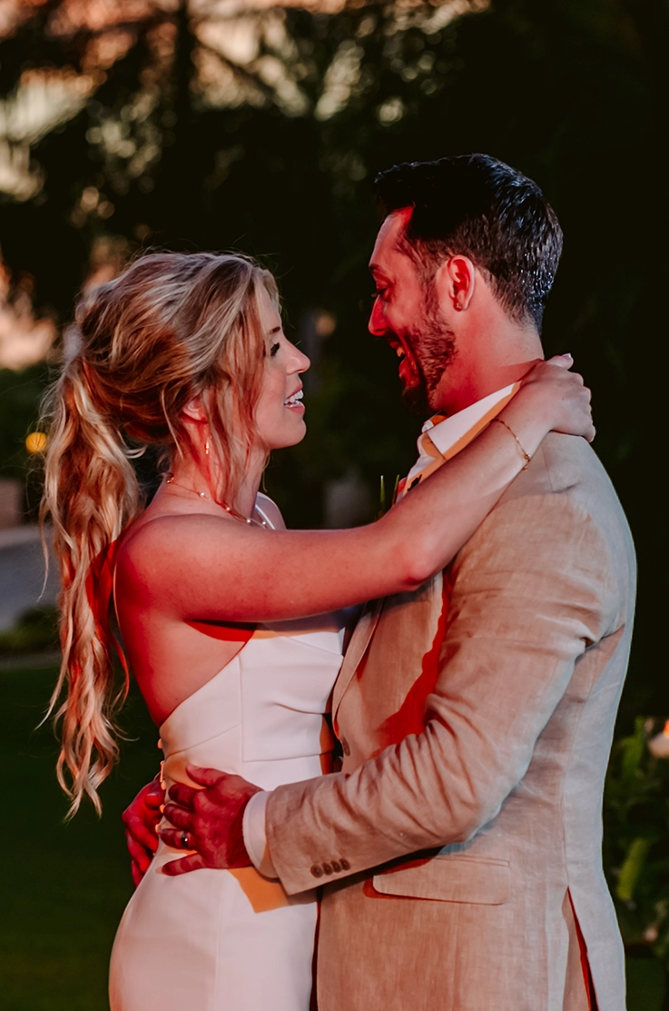 Close up of groom embracing his bride in a crepe fitted gown with buttons and a pleated bodice, near palm trees as the sun sets. 