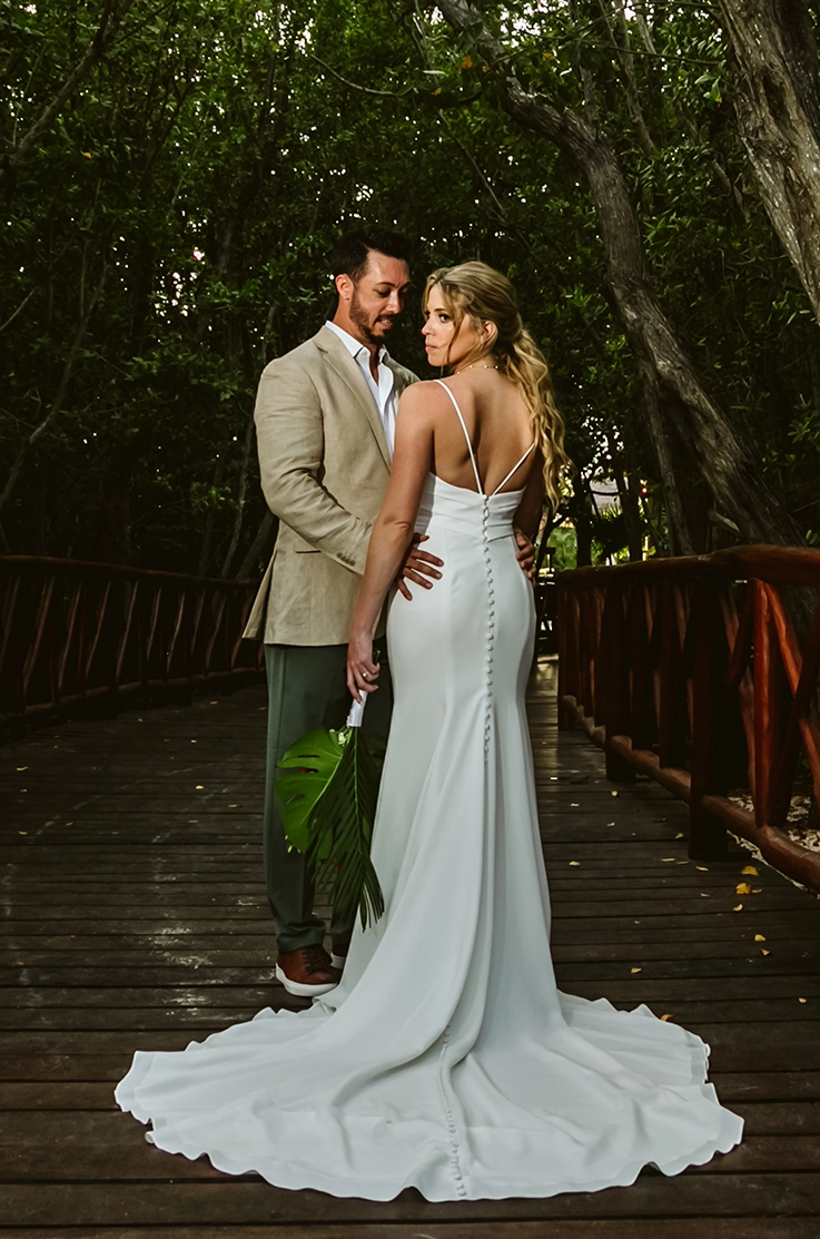 Bride in a fit and flare crepe gown with thin straps and a buttoned back, poses with her groom on a wooden bridge under trees.