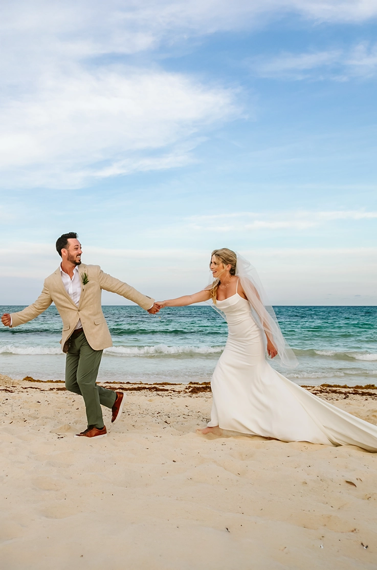 Bride in a pleated crepe bodice gown with fit and flare skirt, and a veil, running on a beach at the shore next to groom. 