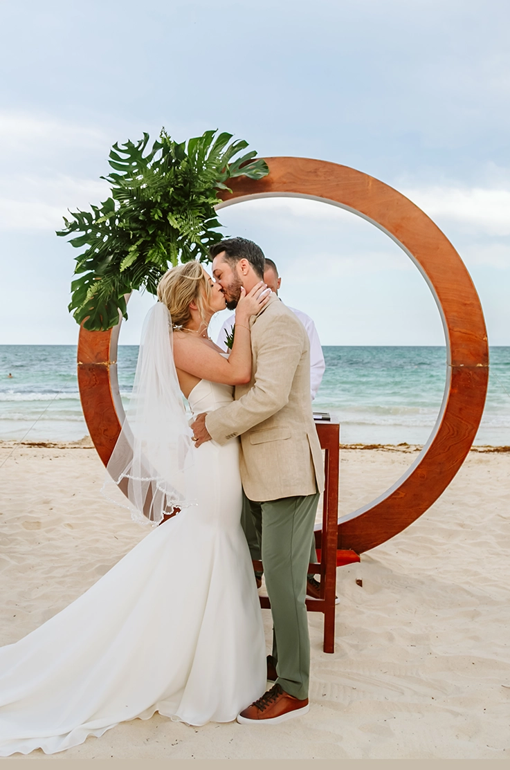 Bride in a crepe princess seamed gown with fit and flare skirt and a veil, kisses groom at a wooden arch on a beach. 