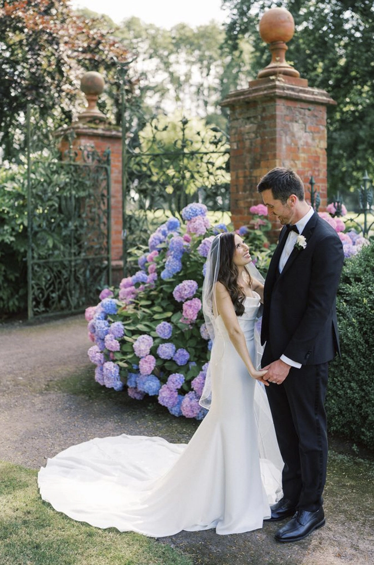 Bride in a crepe pleated bodice gown with a scoop neckline and a veil, looks up at groom by tall garden gates.