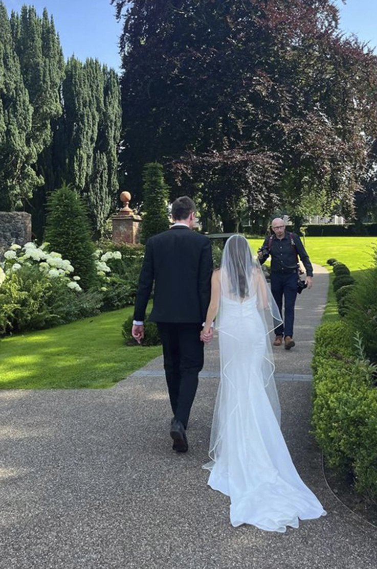 Back of bride in a fit and flare crepe wedding dress with a veil, is walking beside her groom in a manicured garden.
