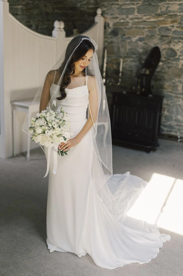 Bride poses in a scoop neck crepe gown with a pleated bodice and a veil, looking to her side and is holding a white bouquet. 