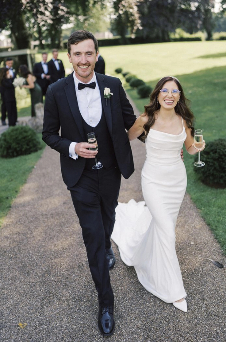 Bride in a pleated crepe bodice gown with a scoop neck, and thin straps, walks alongside her groom, holding champagne glasses .