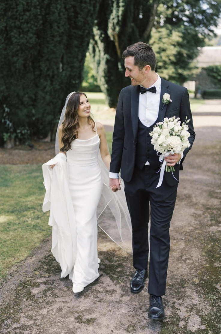 Bride in a scoop neck crepe fitted gown with a veil, holds her train and walks beside the groom on an outdoor path.