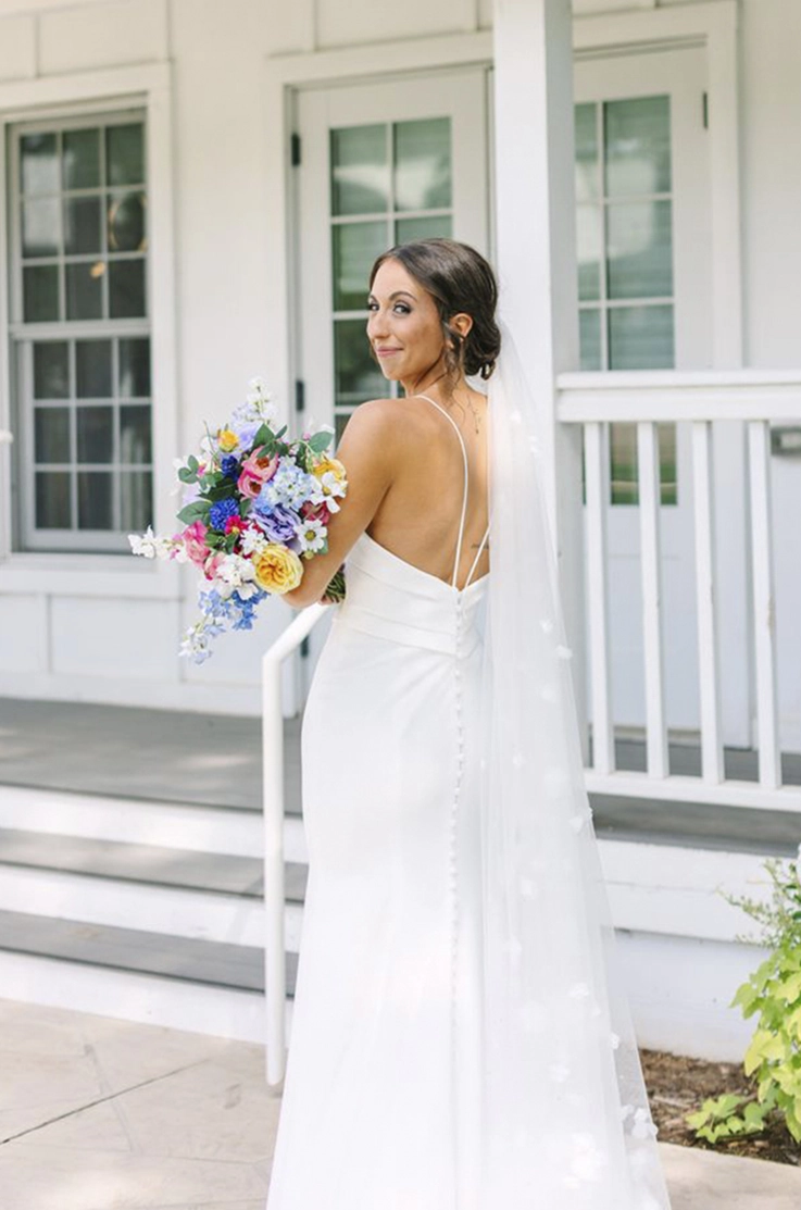 Bride looks over her shoulder in a crepe fit and flare gown with a low buttoned back and veil, in front of the venue doors.