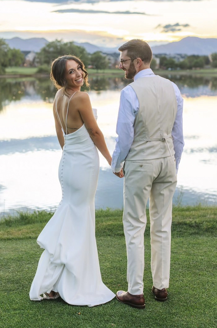Bride poses in a crepe fitted wedding dress with pleating and a low back, standing next to her groom by a lake.
