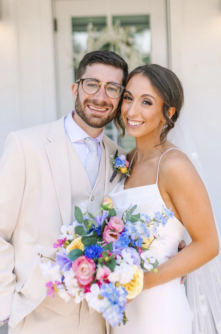 Bride in a sleeveless crepe gown with a scoop pleated bodice and veil, poses next to groom and holds a colorful bouquet.