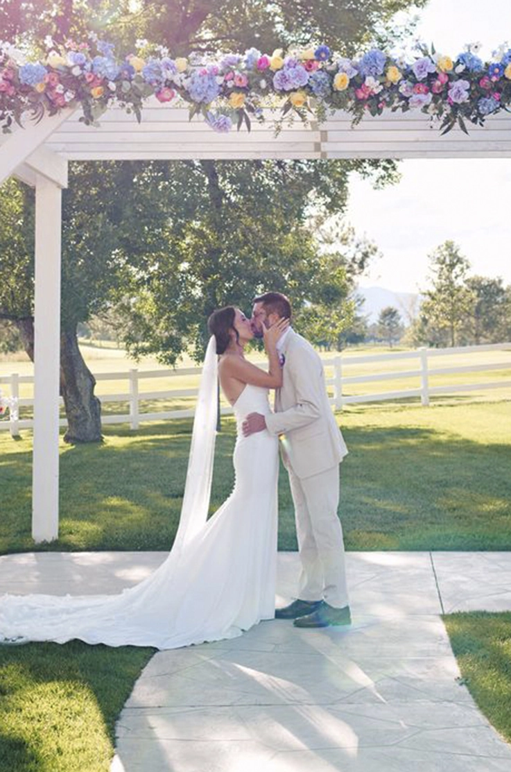 Groom kisses bride in a crepe fit and flare gown with buttons down the back and veil, under a white flower decorated pergola. 