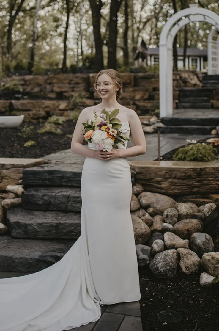 Smiling bride in a fit and flare crepe gown, holding a rustic-colored bouquet and standing on a garden stone path. 