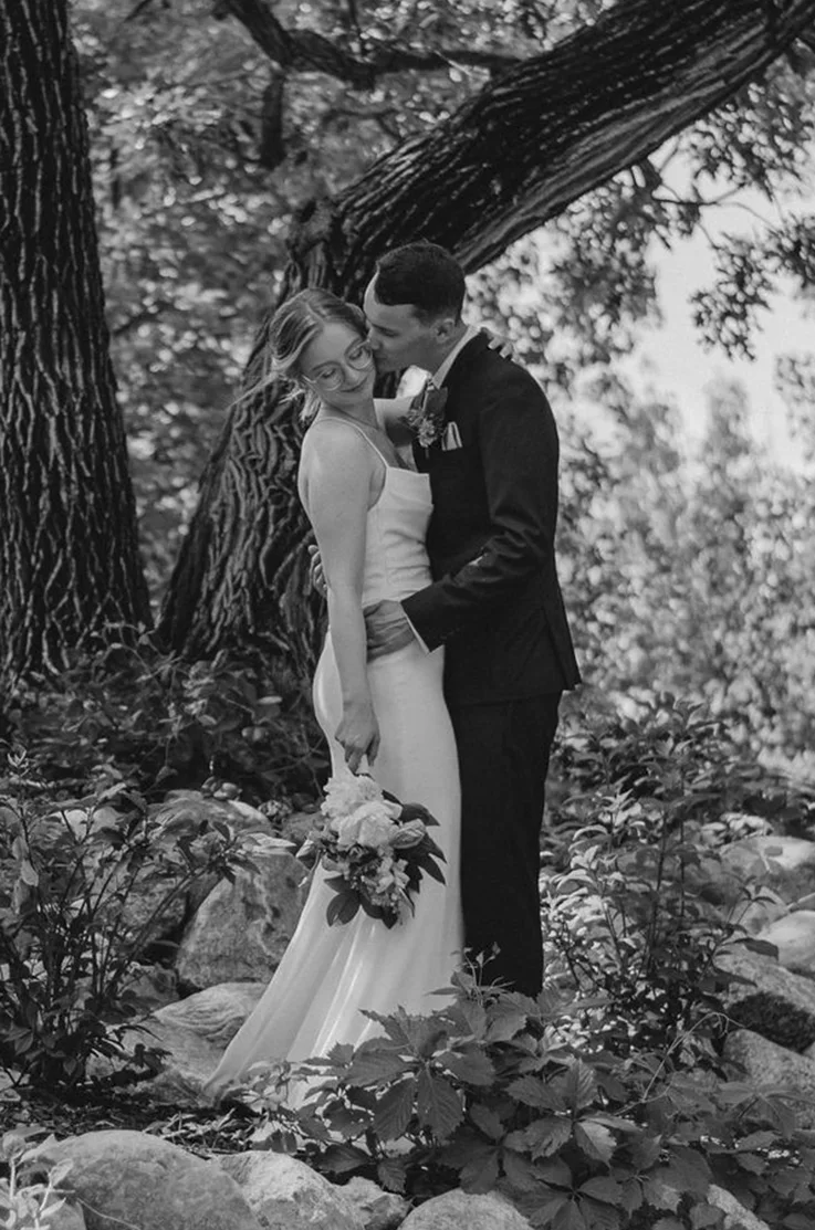 Groom embraces bride in a crepe pleated wedding dress, holding a bouquet under a large tree in black and white.