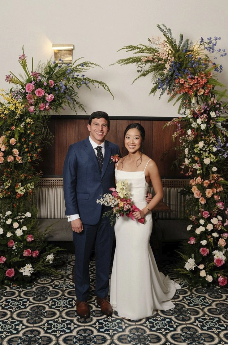 Bride in a crepe fit and flare gown with a scoop neckline, smiles next to groom in a blue suit between floral arches indoors.    