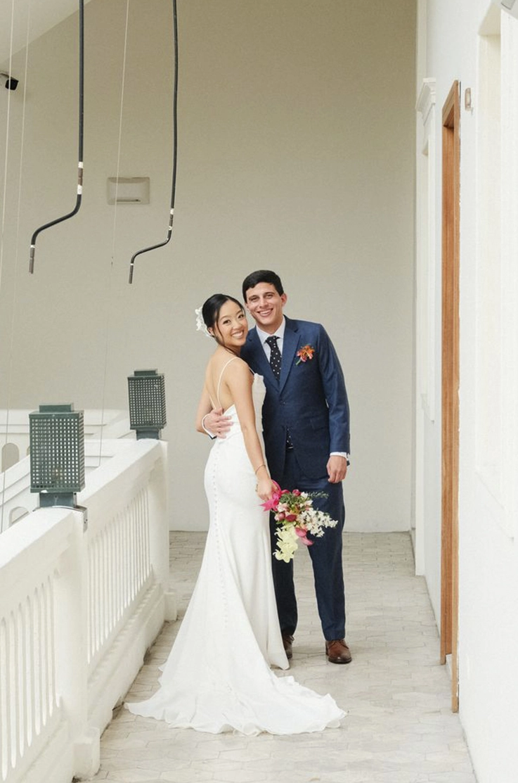 Bride in a spaghetti strap crepe fitted wedding dress, holding a bouquet and poses beside groom by a railing in a corridor.