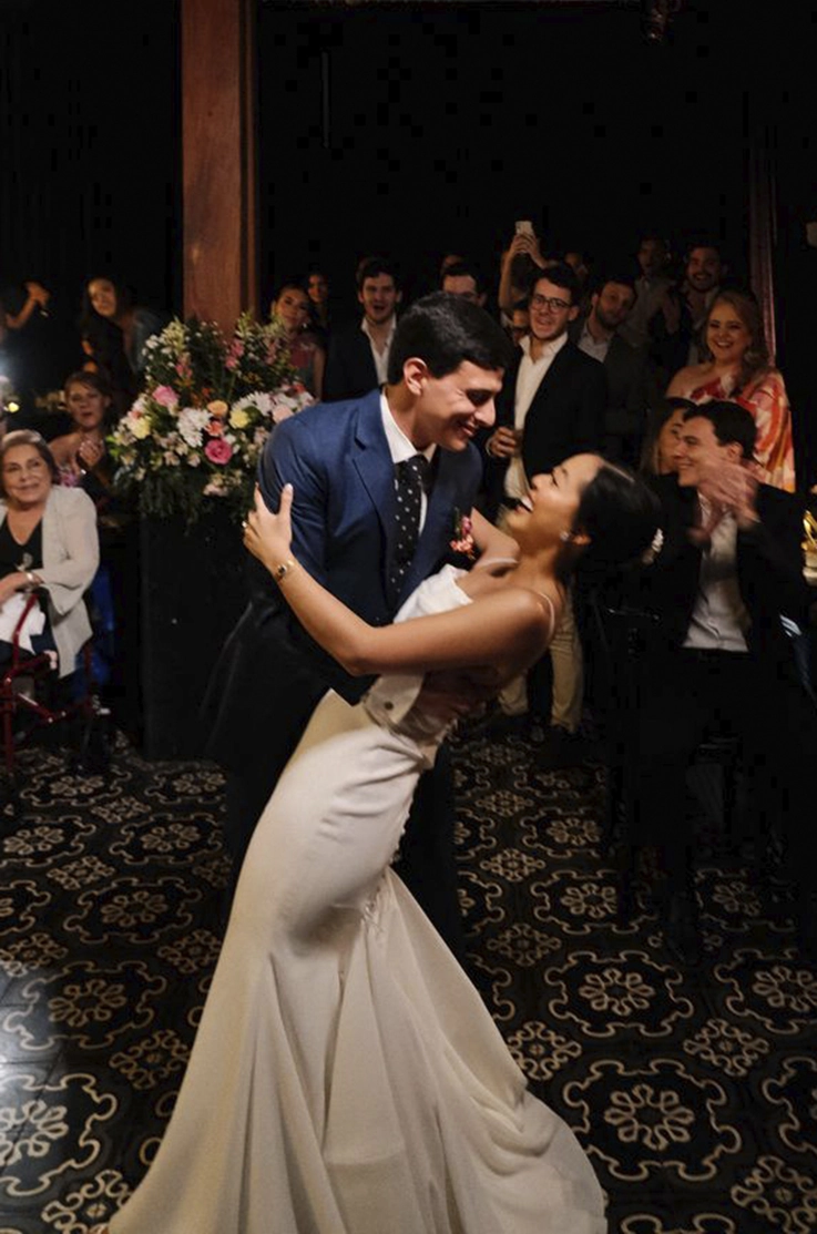 Groom dips his bride in a crepe fit and flare gown with buttons on the back and pleating, in front of guests in blurry photo.