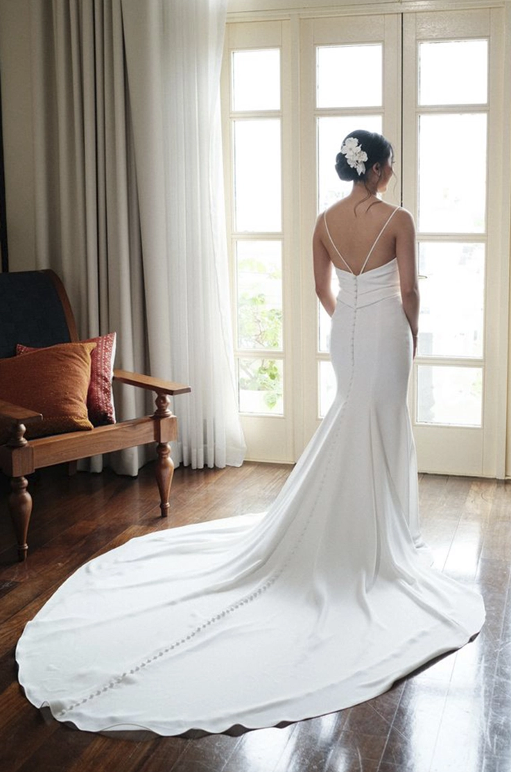 Back of bride looking out a window in a sleeveless fit and flare gown with thin straps and buttons down the train.