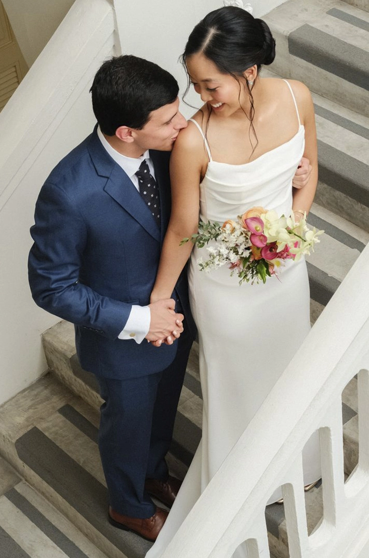 Bride in a pleated crepe bodice gown with spaghetti straps poses with groom on a staircase, taken from above. 