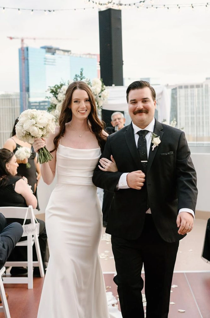 Bride in a crepe fitted wedding dress with pleating, walks alongside her groom down the aisle on a city rooftop.