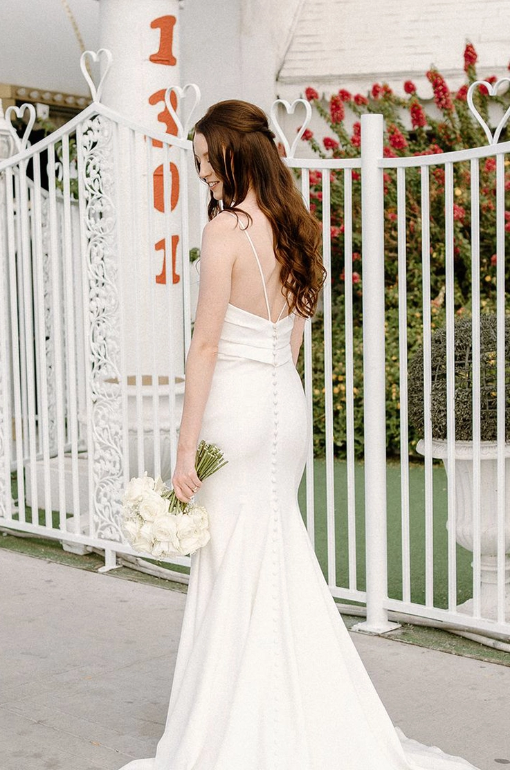 Back of bride posing in a fit and flare crepe gown with a low back and button detail, standing at a garden gate.