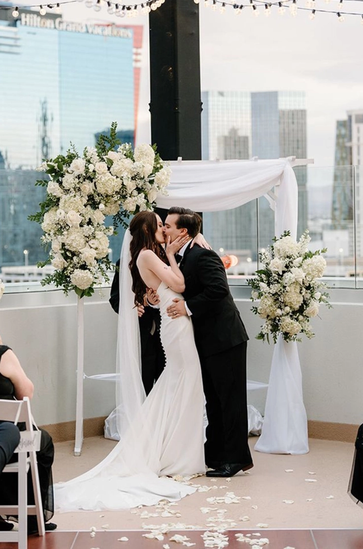 Groom kisses his bride in a fit and flare crepe gown with buttons and a veil, at floral arch on a rooftop.