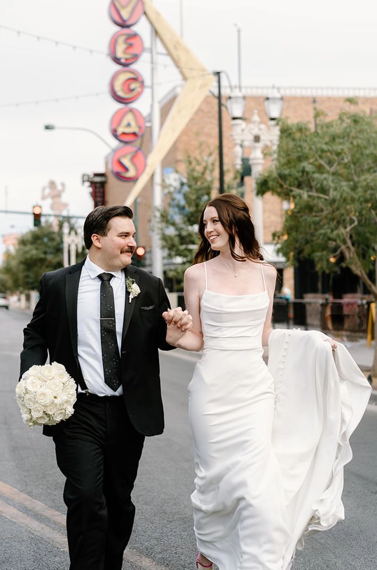 Bride in a crepe pleated bodice gown, holds her train while walking next to the groom, in front of a Vegas sign.