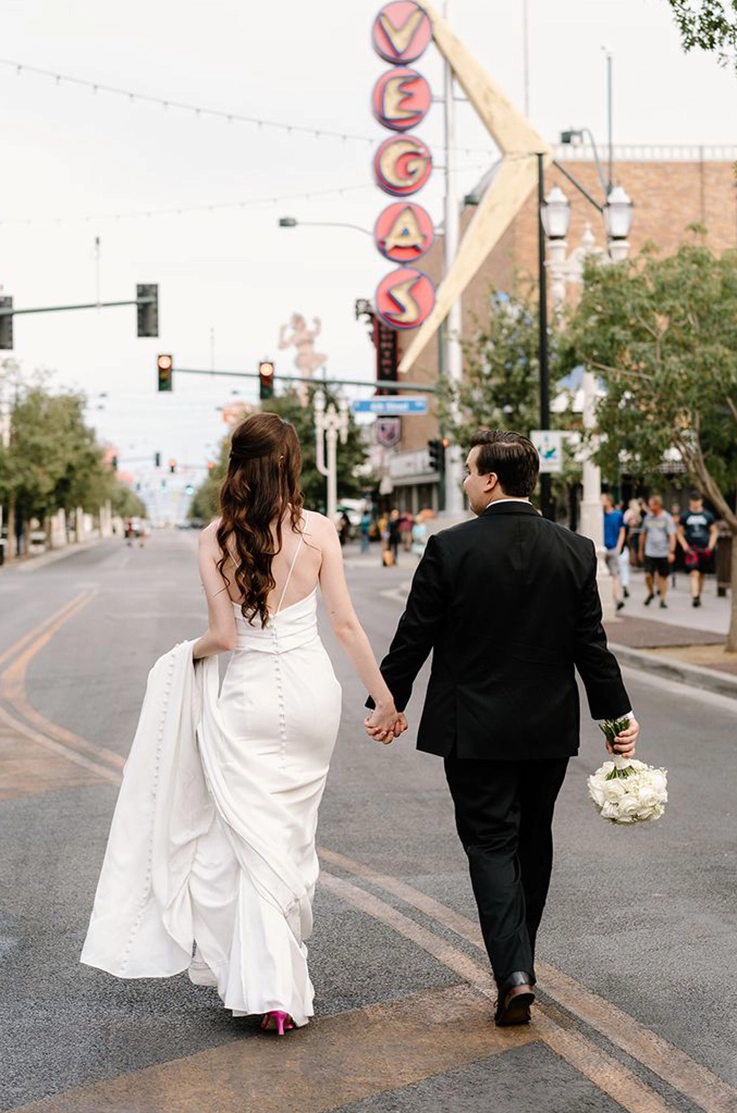 Bride in a crepe spaghetti strap gown with buttons on the back, walks hand in hand with her groom down a city street.