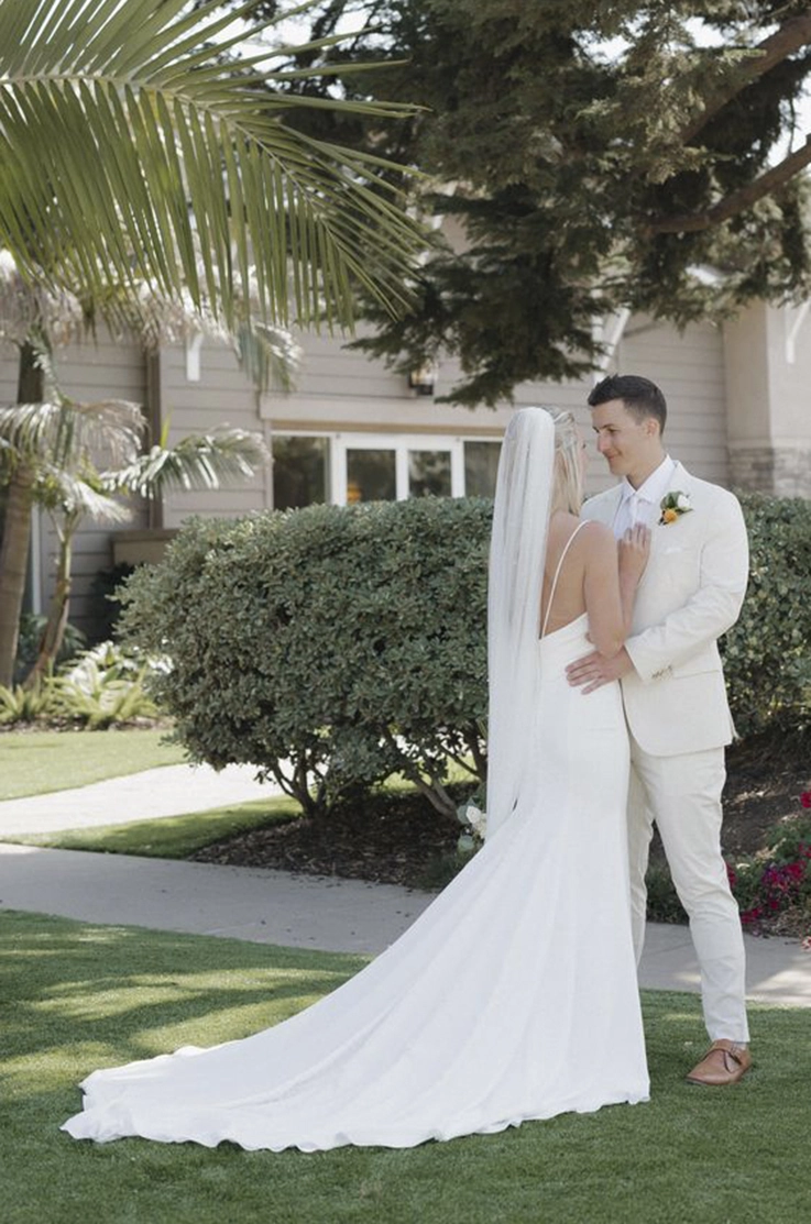 Groom embraces his bride in a sleeveless crepe fitted gown with a veil, standing beneath palm trees on a green lawn.