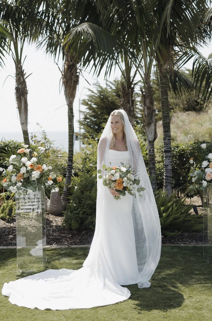 Bride in a crepe fit and flare gown with a scoop neckline and a veil, is holding a bouquet between flowers and palm trees. 