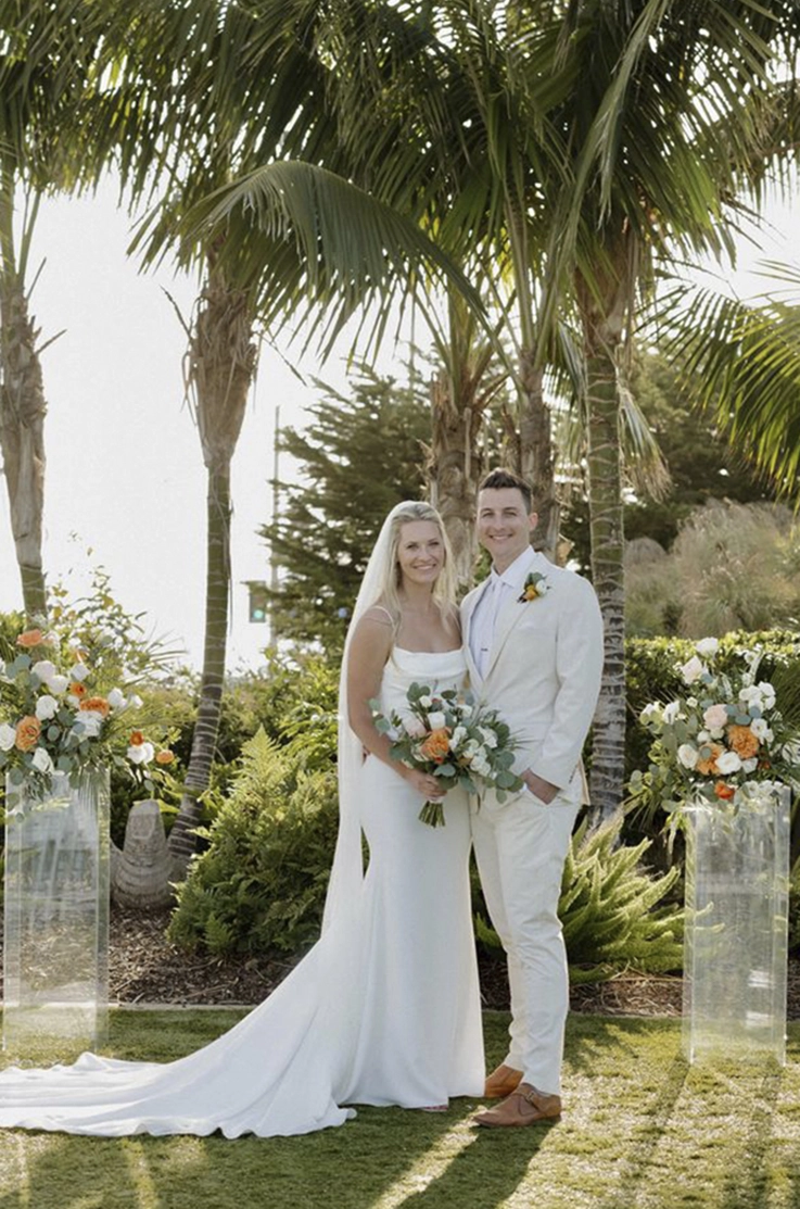 Bride in a crepe fit and flare gown with a scoop neck and veil, poses beside groom between flower posts under palm trees.