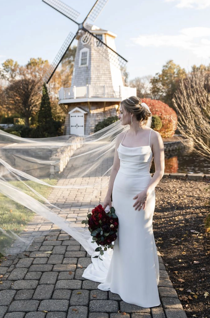 Bride poses by a windmill in a scoop neck crepe gown with fit and flare skirt and a flowing veil, holding a bouquet. 