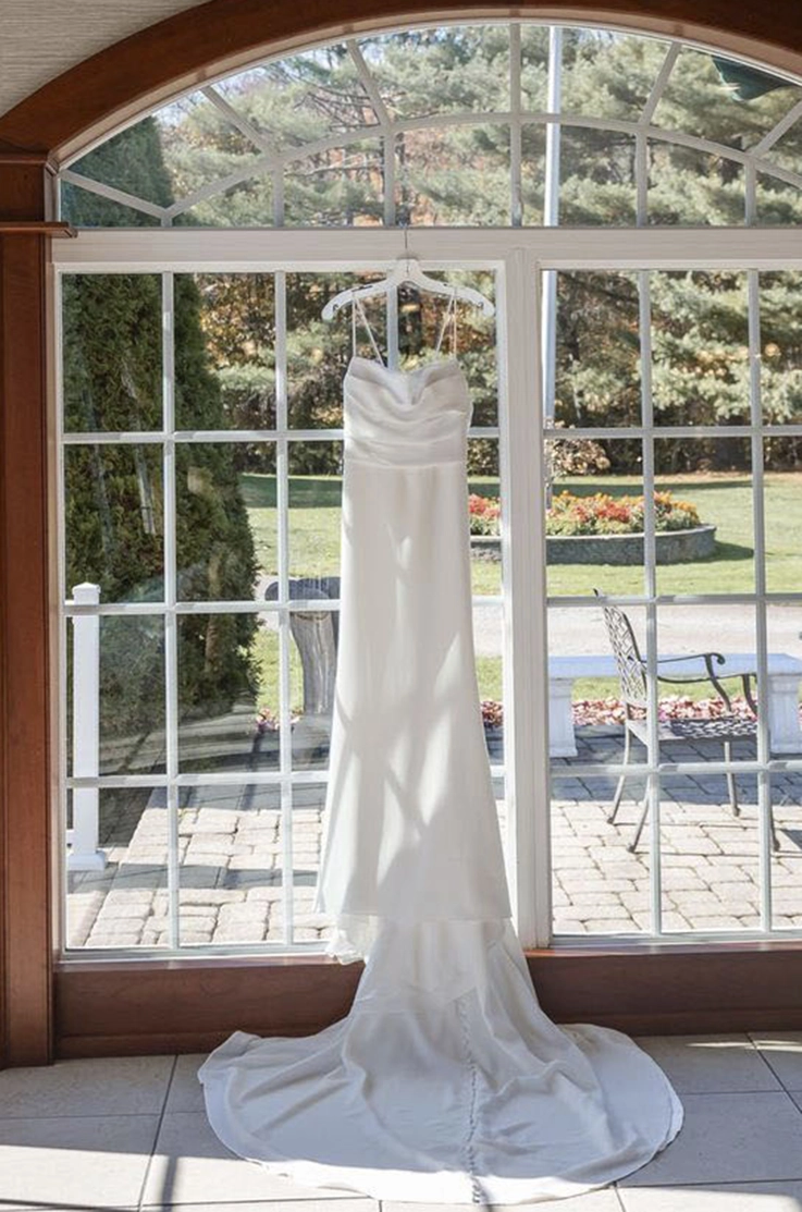 A spaghetti strap crepe wedding dress with a pleated bodice and buttons, hangs on a tall window facing a sunny garden.