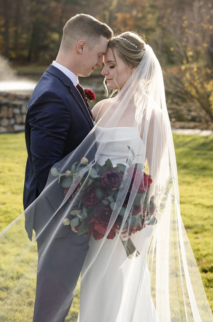 Groom embraces bride in a pleated crepe bodice gown with a veil and dark red bouquet, standing on a sunny lawn