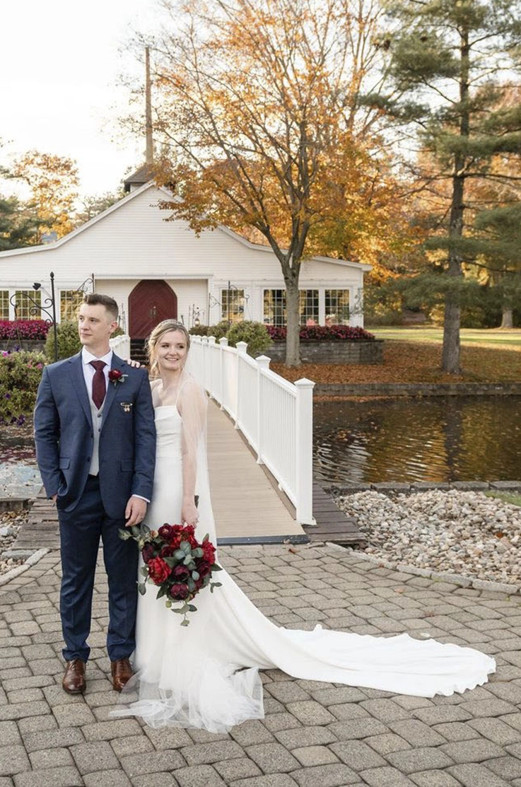 Bride in a crepe pleated gown with a veil, poses next to her groom on a stone pathway in front of a white bridge and river. 