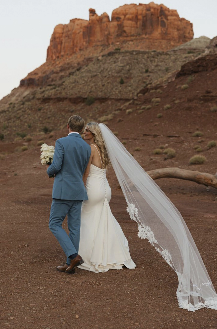 Bride in a fit and flare pleated gown with a flowing veil, walks next to her groom at a red rock canyon. 