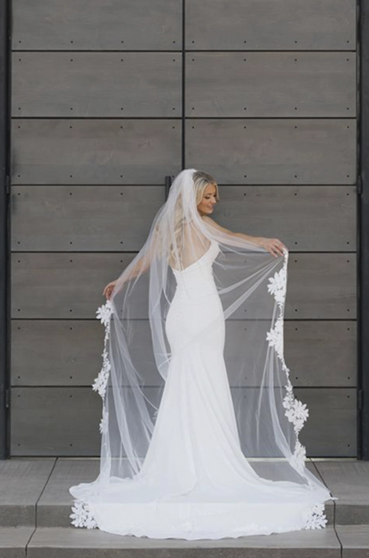 Bride poses in a crepe fit and flare wedding dress with a scoop neckline, holding up a lace-trim veil by tall venue doors.