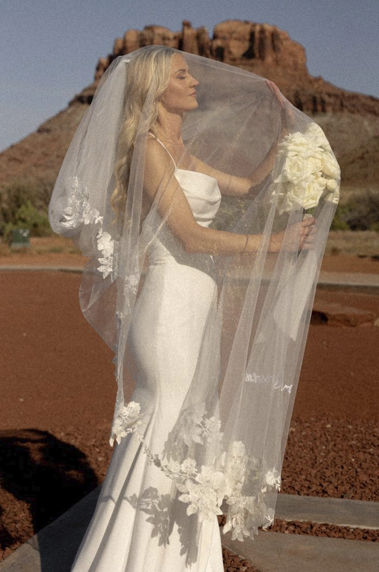Bride poses under a lace-trim veil in a scoop neck crepe gown with a fit and flare skirt, and is holding a white bouquet in a red canyon.